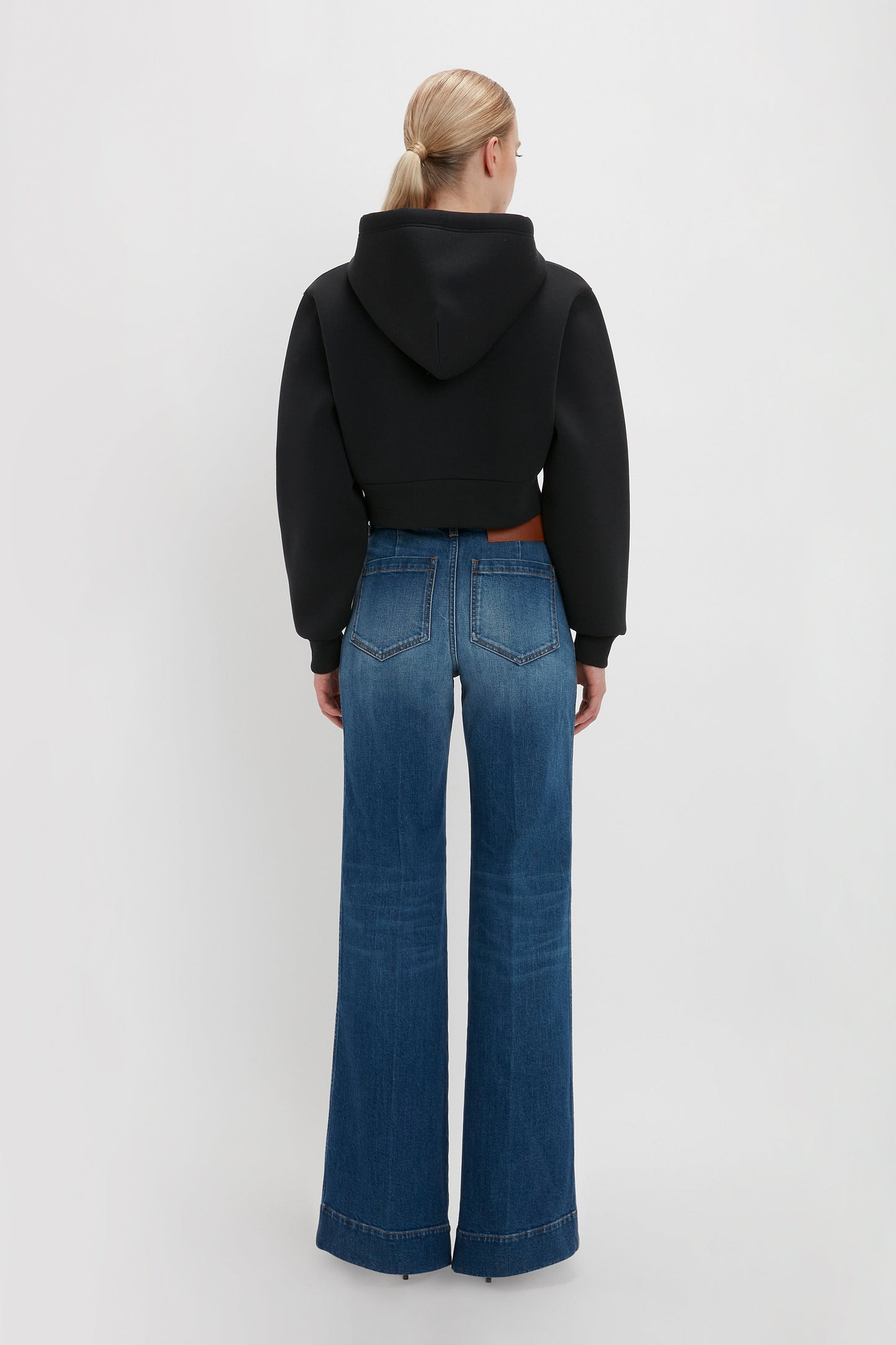 A person with blonde hair is standing with their back to the camera, wearing a Victoria Beckham Cropped Neoprene Hoodie In Black and wide-leg blue jeans.