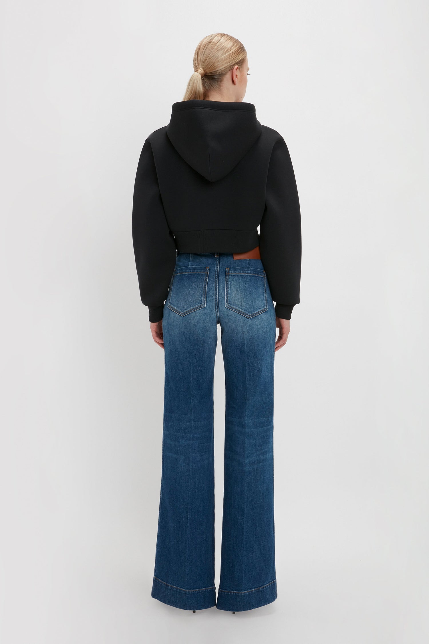 A person with blonde hair is standing with their back to the camera, wearing a Victoria Beckham Cropped Neoprene Hoodie In Black and wide-leg blue jeans.