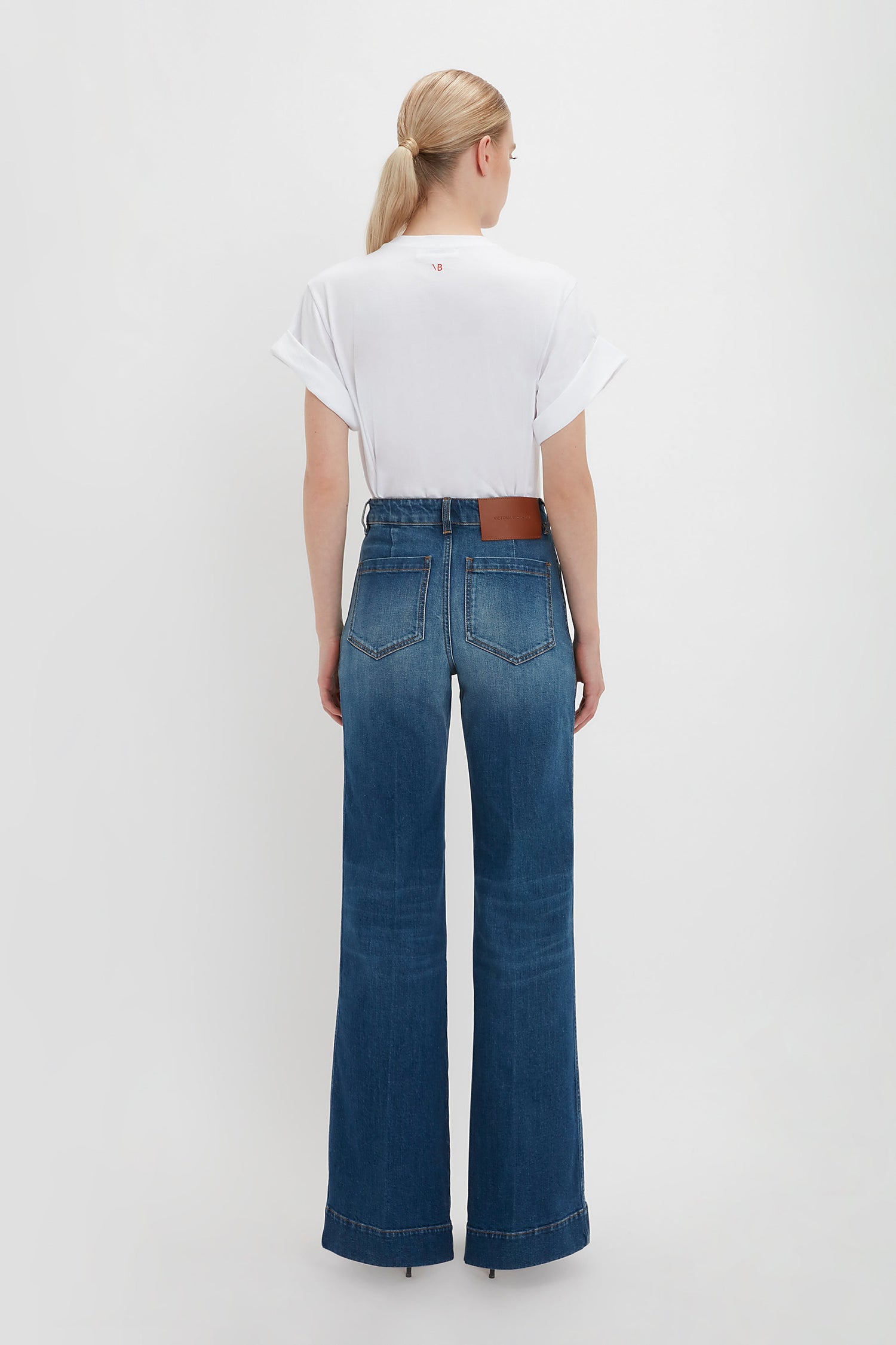 A person with blond hair in a ponytail, wearing an oversized Victoria Beckham Asymmetric Relaxed Fit T-Shirt In White and blue high-waisted wide-leg jeans with asymmetric folded sleeves, stands facing away.