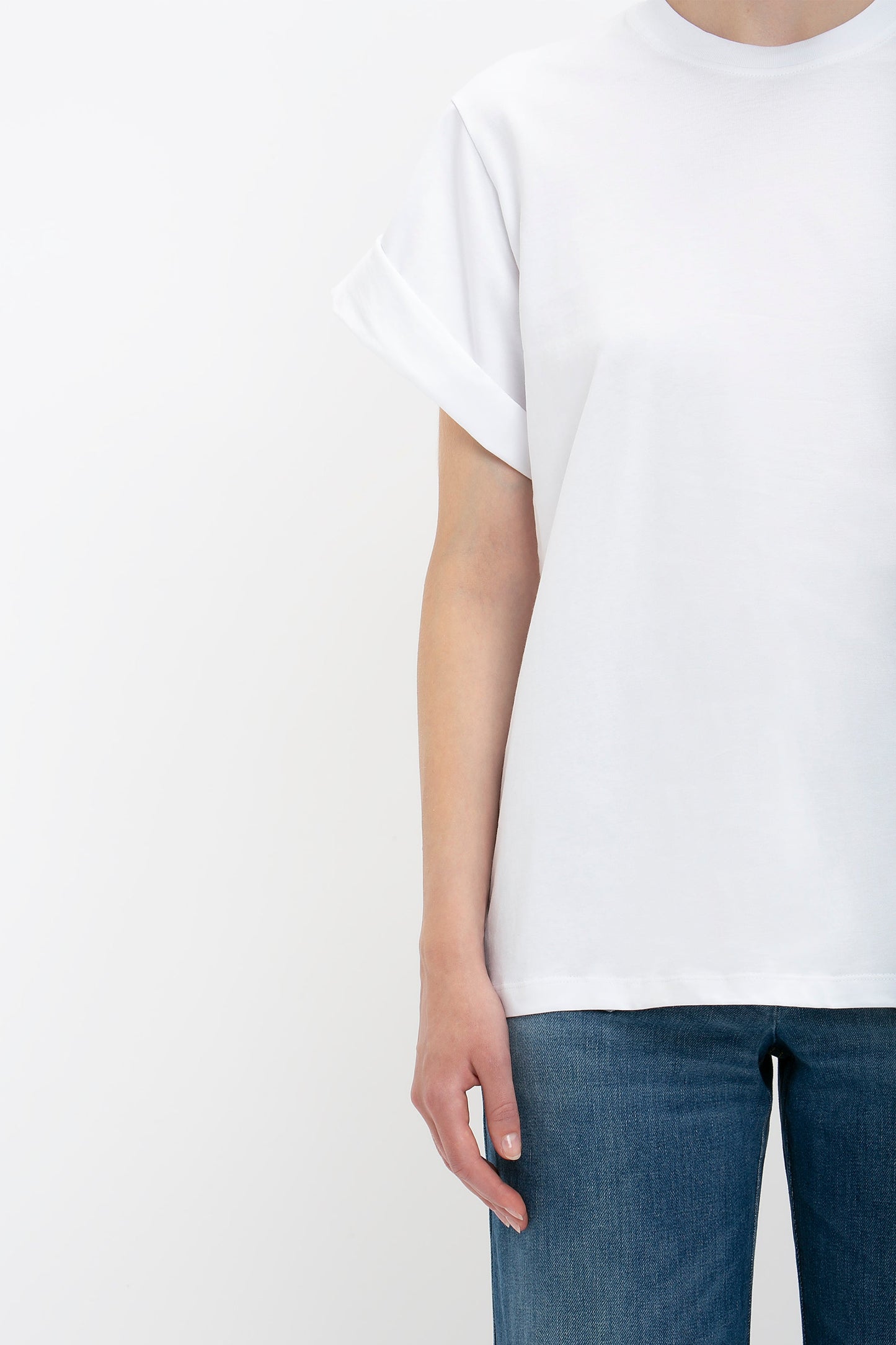 Person in an oversized Victoria Beckham Asymmetric Relaxed Fit T-Shirt In White with folded sleeves and blue jeans, standing against a blank white background. Only the upper body is visible.