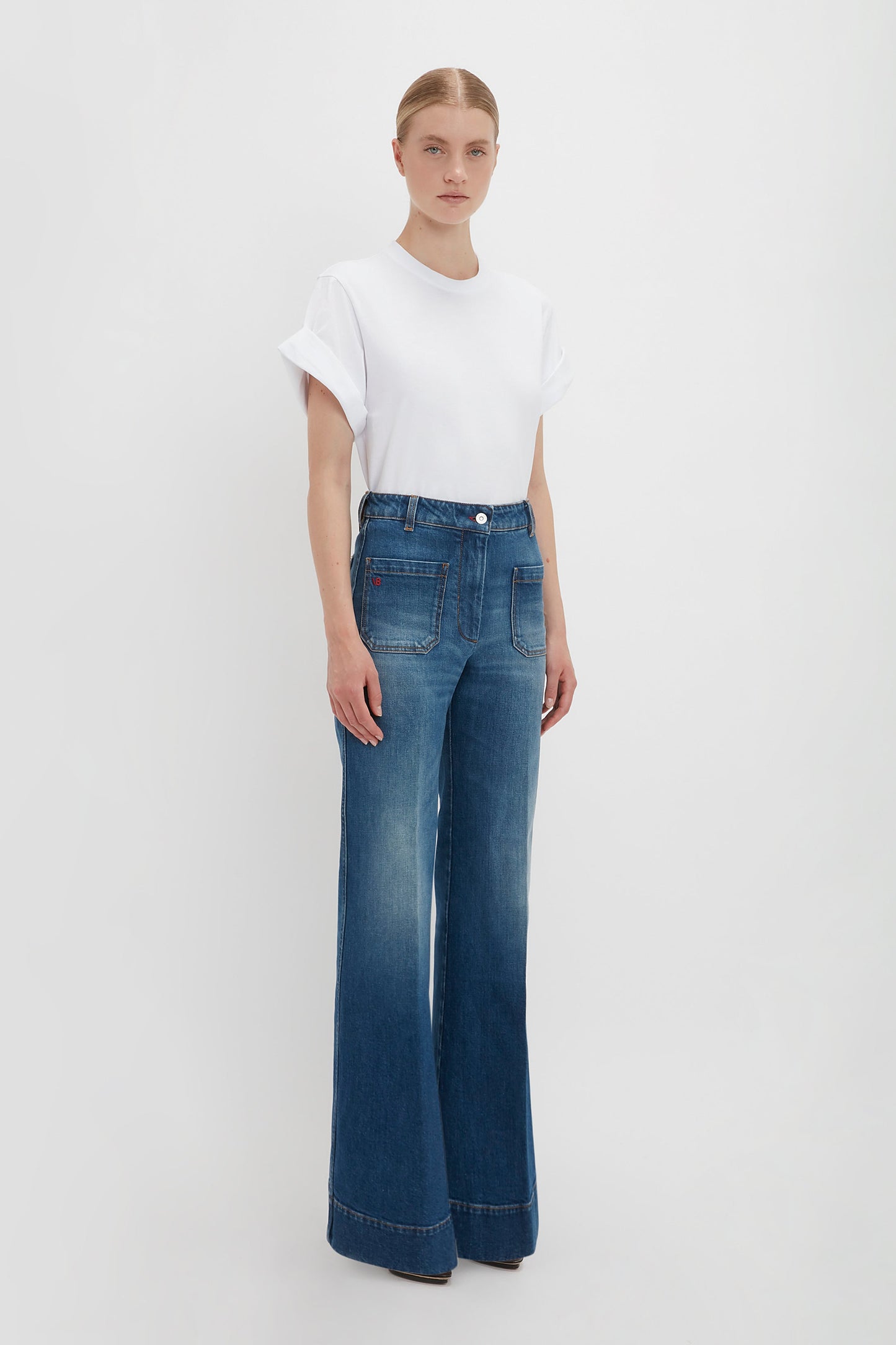 A person stands against a plain white background, wearing an oversized Asymmetric Relaxed Fit T-Shirt In White by Victoria Beckham and high-waisted, wide-leg blue jeans.